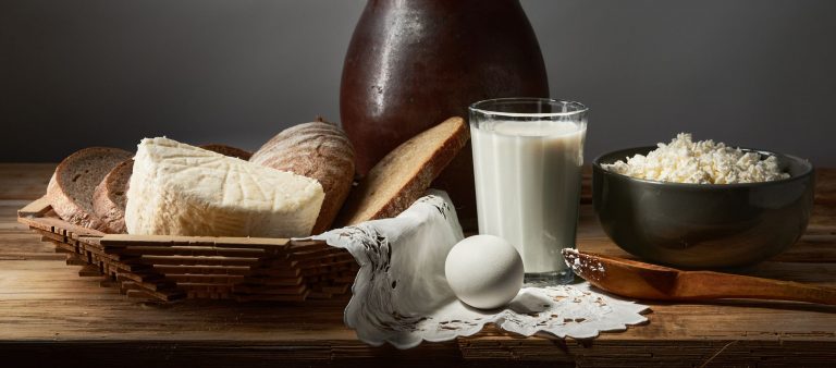 A rustic still life featuring bread, cheese, egg, milk, and cottage cheese on a wooden table.